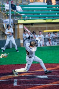 A baseball player swings at a pitch on a colorful field, with teammates cheering from the dugout behind him.