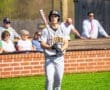 High school baseball player in gray uniform preparing to bat, with spectators in the background at a sunny game.