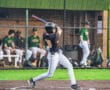A young baseball player swings a bat on the field, with teammates watching from the dugout behind him.