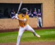 A baseball player swings a bat at home plate, with teammates watching from the dugout in the background.