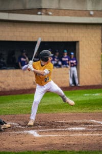 A baseball player swings a bat at home plate, with teammates watching from the dugout in the background.