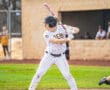 High school baseball player in striped uniform prepares to bat on a sunny field with spectators in the background.