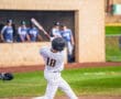 A baseball player in striped uniform swings a bat, preparing to hit the ball during a game with teammates in the background.