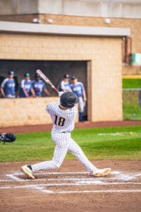 A baseball player in striped uniform swings a bat, preparing to hit the ball during a game with teammates in the background.