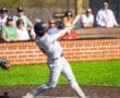 Baseball player swings bat at home plate during game, with fans watching in the background on a sunny day.