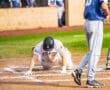 Baseball player on the ground after sliding into home plate, with teammates watching from the dugout in the background.