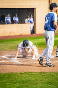 Baseball player on the ground after sliding into home plate, with teammates watching from the dugout in the background.