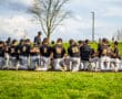 Baseball team kneeling on field, listening to coach during practice on a sunny day with clouds in the background.
