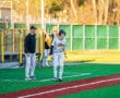Baseball players on field during practice, coach guiding a player at third base in a sunny, vibrant setting.