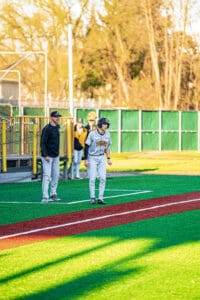 Baseball players on field during practice, coach guiding a player at third base in a sunny, vibrant setting.