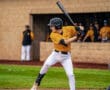 A young baseball player swings a bat at home plate during a game, with teammates in the dugout in the background.