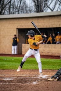 A young baseball player swings a bat at home plate during a game, with teammates in the dugout in the background.