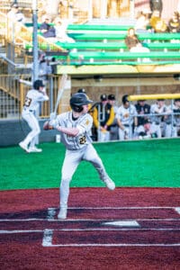 A young baseball player stands ready to bat, with teammates and spectators in the background at a lively game.