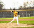 Baseball pitcher in a yellow jersey delivering a pitch on the mound during a game, with a grassy field backdrop.