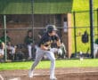 A baseball player in a black jersey runs towards first base on a sunny day, with teammates in the dugout behind him.