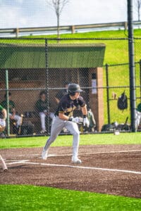 A baseball player in a black jersey runs towards first base on a sunny day, with teammates in the dugout behind him.