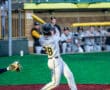 A baseball player swings a bat on the field, while teammates watch from the dugout in the background.
