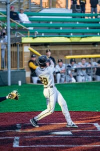 A baseball player swings a bat on the field, while teammates watch from the dugout in the background.