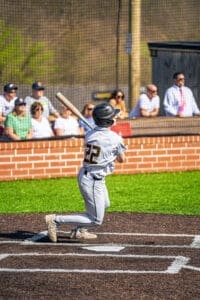 Young baseball player in gray uniform swings bat during sunny game, with spectators in background enjoying the match.