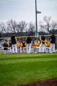 Baseball team gathers for a motivational talk on the field, wearing yellow jerseys and black jackets during practice.