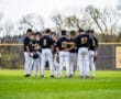 A baseball team huddles in the outfield, discussing strategy on a sunny day. Team members wear black and white uniforms.