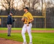 Baseball player in yellow jersey stands on the field, focused, as a coach gestures in the background during practice.