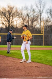 Baseball player in yellow jersey stands on the field, focused, as a coach gestures in the background during practice.