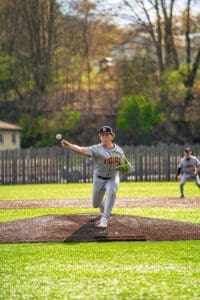 Young baseball pitcher in gray uniform throws a pitch on a sunny day, with teammates in action on the field behind him.