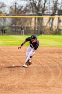 A player in a black jersey and white pants fields a ground ball on a sunlit baseball diamond.