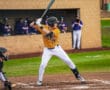 A baseball player in an orange jersey prepares to bat during a game, with teammates visible in the background.
