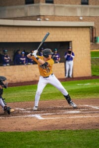 A baseball player in an orange jersey prepares to bat during a game, with teammates visible in the background.