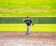 A young baseball player dressed in a black jersey throws the ball on a sunny day at the baseball field.