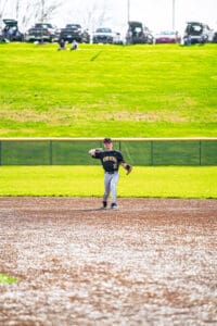 A young baseball player dressed in a black jersey throws the ball on a sunny day at the baseball field.