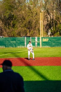 Baseball player in white uniform on the field, ready to play, with a vibrant green and red turf background.