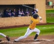 A baseball player in a yellow jersey swings a bat during a game, with teammates watching from the dugout.