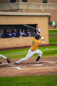 A baseball player in a yellow jersey swings a bat during a game, with teammates watching from the dugout.
