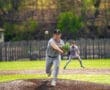 A young baseball pitcher delivers a fastball on a sunny day, with a teammate positioned in the background.