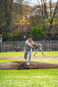 A young baseball pitcher delivers a fastball on a sunny day, with a teammate positioned in the background.