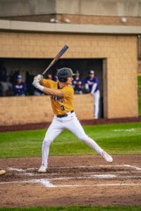 Player in a yellow baseball jersey swings a bat on the field, with teammates visible in the background.