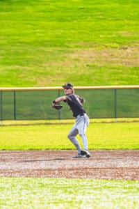 A young baseball player prepares to throw, wearing a black jersey and cap, on a sunny day at the field.