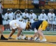 Two lacrosse players face off for the ball, with a referee and teammates in the background on a vibrant field.