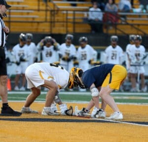 Two lacrosse players face off for the ball, with a referee and teammates in the background on a vibrant field.