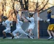 High school lacrosse players in action as a goalie defends against a shot during a sunset game.
