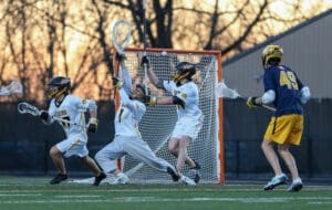 High school lacrosse players in action as a goalie defends against a shot during a sunset game.