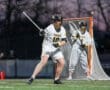 Lacrosse players in action at dusk, one preparing to pass while the goalie stands ready behind the net.
