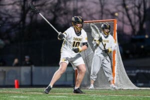 Lacrosse players in action at dusk, one preparing to pass while the goalie stands ready behind the net.