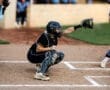 Girl catcher in black uniform prepares to receive a pitch in a softball game, with umpire and players in the background.