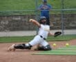 A catcher in a white uniform dives to catch a ball during a softball game, with tennis balls scattered nearby.