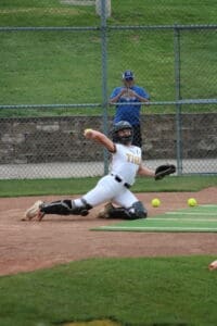 A catcher in a white uniform dives to catch a ball during a softball game, with tennis balls scattered nearby.