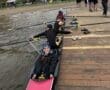 Rowing team prepares for practice on a cloudy day, with team members and equipment on the water's edge.
